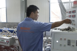 A person wearing a blue shirt operates a control panel in an industrial setting. The room features various machinery and control units with buttons and displays. Natural light enters through the windows, illuminating the workspace.