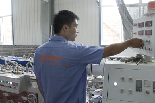 A person wearing a blue shirt operates a control panel in an industrial setting. The room features various machinery and control units with buttons and displays. Natural light enters through the windows, illuminating the workspace.