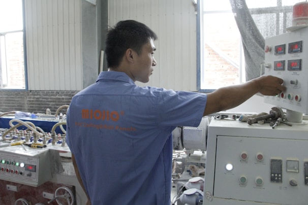 A person wearing a blue shirt operates a control panel in an industrial setting. The room features various machinery and control units with buttons and displays. Natural light enters through the windows, illuminating the workspace.
