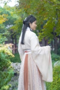 A woman wearing traditional clothing stands gracefully in a serene garden. Her long, flowing robe features light and earthy tones, complemented by intricate lace details. She holds a slender black object, possibly a fan, as she gazes into the lush, green surroundings.