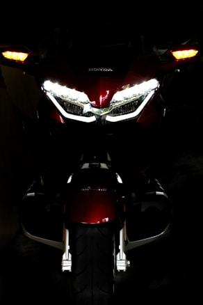 A motorcycle is viewed from the front in a dark environment, highlighting its headlights and turn signals. The brand logo is visible above the headlights on the maroon body of the bike.