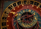 An intricate astronomical clock featuring Roman numerals, zodiac signs, and detailed craftsmanship. The face has concentric rings with illustrations representing different zodiac symbols and medieval Gothic lettering. The color scheme includes gold, black, red, and teal, centering around a complex design with movable components.
