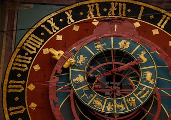 An intricate astronomical clock featuring Roman numerals, zodiac signs, and detailed craftsmanship. The face has concentric rings with illustrations representing different zodiac symbols and medieval Gothic lettering. The color scheme includes gold, black, red, and teal, centering around a complex design with movable components.