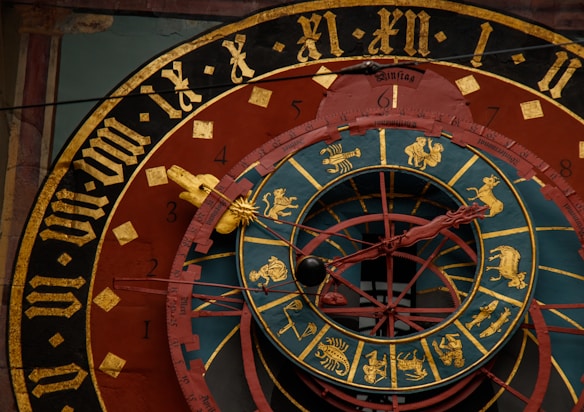 An intricate astronomical clock featuring Roman numerals, zodiac signs, and detailed craftsmanship. The face has concentric rings with illustrations representing different zodiac symbols and medieval Gothic lettering. The color scheme includes gold, black, red, and teal, centering around a complex design with movable components.