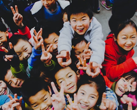A group of children enthusiastically crowd together, many of them smiling and flashing peace signs with their hands. They appear to be dressed warmly in colorful clothing, suggesting a lively and cheerful atmosphere.