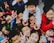 A group of children enthusiastically crowd together, many of them smiling and flashing peace signs with their hands. They appear to be dressed warmly in colorful clothing, suggesting a lively and cheerful atmosphere.