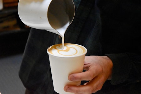 A person is pouring steamed milk into a cup of coffee, forming an intricate latte art pattern on top. The action is captured close-up, focusing on the swirling design and the contrast between the coffee and milk.