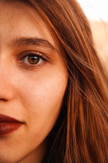 A close-up of a person's face featuring one eye, eyebrow, a portion of the nose, and lips. The person has brown hair and brown eyes, with light makeup on the lips.