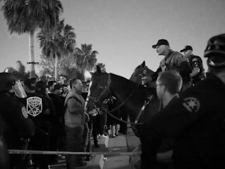 A black and white photo showing a group of police officers on horseback facing a crowd of people. The scene appears to be tense, with a confrontation between the officers and the crowd. Palm trees can be seen in the background, suggesting a possible urban or suburban setting.