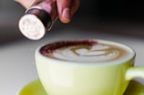A close-up of a cappuccino in a light green cup with beautiful latte art on top. A hand is sprinkling cocoa or cinnamon powder from a shaker onto the frothy surface.