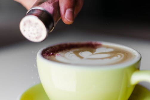 A close-up of a cappuccino in a light green cup with beautiful latte art on top. A hand is sprinkling cocoa or cinnamon powder from a shaker onto the frothy surface.