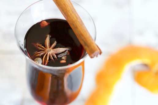 A glass contains a dark liquid, topped with a star anise and pieces of what appear to be spices. A cinnamon stick rests inside the glass, adding to the seasonal, aromatic theme. In the background, an orange peel adds a splash of color, complementing the warm tones of the drink.