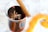 A glass contains a dark liquid, topped with a star anise and pieces of what appear to be spices. A cinnamon stick rests inside the glass, adding to the seasonal, aromatic theme. In the background, an orange peel adds a splash of color, complementing the warm tones of the drink.