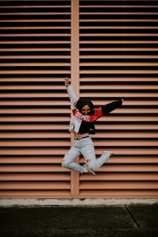 A person is captured mid-air in a joyous jump in front of a vertically striped peach-colored wall. They wear a color-blocked sweater and ripped jeans, exuding a sense of freedom and energy.