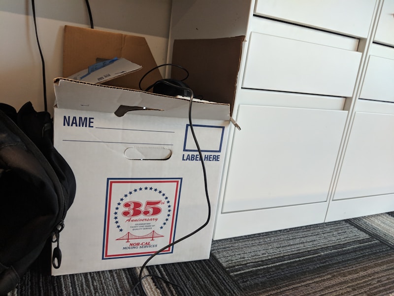 A cardboard box labeled for moving services is placed on a carpeted floor. The box has a design indicating a 35th anniversary. There is a cable draped over the box, and a backpack is partially visible next to it. Nearby, there are white drawers.