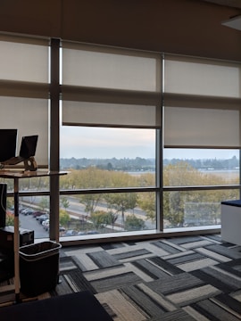 An office interior with large windows allowing a view of an outdoor scene with trees and a roadway. The room features a standing desk with a laptop and a trash bin nearby. The floor is covered in a patterned carpet with gray and black tones.