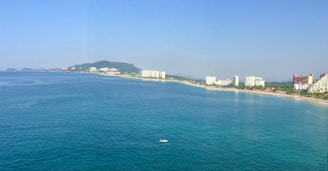 A coastal cityscape featuring a long stretch of beach bordered by a calm blue sea. High-rise buildings and resorts line the shore, with a backdrop of green hills in the distance. A small boat is visible on the water, adding a sense of scale to the vast ocean.