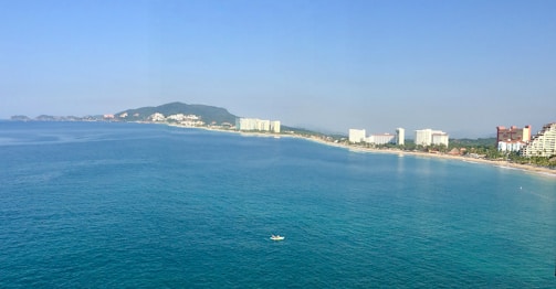 A coastal cityscape featuring a long stretch of beach bordered by a calm blue sea. High-rise buildings and resorts line the shore, with a backdrop of green hills in the distance. A small boat is visible on the water, adding a sense of scale to the vast ocean.