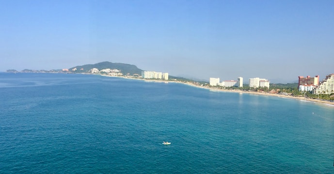 A coastal cityscape featuring a long stretch of beach bordered by a calm blue sea. High-rise buildings and resorts line the shore, with a backdrop of green hills in the distance. A small boat is visible on the water, adding a sense of scale to the vast ocean.