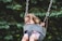 A young child with windblown hair sits on a swing set secured with a safety seat. The child appears to be enjoying the swing, surrounded by blurred greenery in the background.