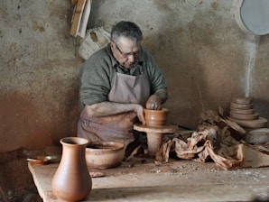 A person is engaged in pottery-making in a rustic workshop. Wearing an apron, the individual is focused on shaping a clay pot on a pottery wheel. The surroundings include a wooden table scattered with pottery tools and partially finished pots. The walls are bare and textured, adding to the earthy feel of the environment.
