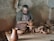A person is engaged in pottery-making in a rustic workshop. Wearing an apron, the individual is focused on shaping a clay pot on a pottery wheel. The surroundings include a wooden table scattered with pottery tools and partially finished pots. The walls are bare and textured, adding to the earthy feel of the environment.