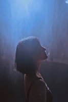 A woman with short dark hair stands under a gentle shower of rain, illuminated by a soft, ethereal blue light creating a serene atmosphere around her.