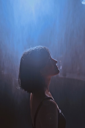 A woman with short dark hair stands under a gentle shower of rain, illuminated by a soft, ethereal blue light creating a serene atmosphere around her.