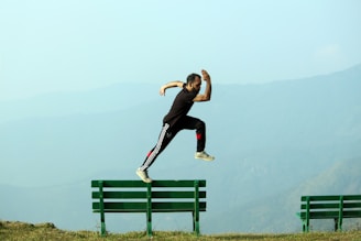 A person is captured in mid-air while jumping from one green bench to another against a backdrop of misty mountains. The individual is dressed in black athletic wear with white and red accents.