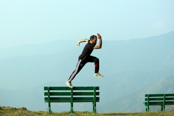 A person is captured in mid-air while jumping from one green bench to another against a backdrop of misty mountains. The individual is dressed in black athletic wear with white and red accents.