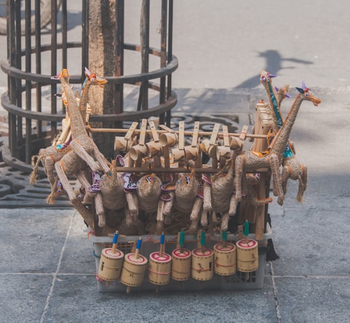 Handcrafted toys made of natural materials such as wood and straw are arranged on a street. The toys include colorful giraffe figures and cylindrical percussion instruments, displayed on a stone sidewalk near a tree.
