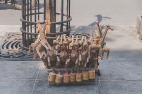 Handcrafted toys made of natural materials such as wood and straw are arranged on a street. The toys include colorful giraffe figures and cylindrical percussion instruments, displayed on a stone sidewalk near a tree.