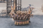 Handcrafted toys made of natural materials such as wood and straw are arranged on a street. The toys include colorful giraffe figures and cylindrical percussion instruments, displayed on a stone sidewalk near a tree.