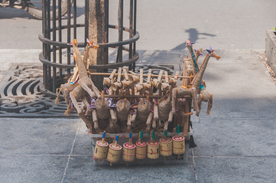 Handcrafted toys made of natural materials such as wood and straw are arranged on a street. The toys include colorful giraffe figures and cylindrical percussion instruments, displayed on a stone sidewalk near a tree.
