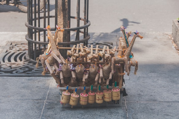 Handcrafted toys made of natural materials such as wood and straw are arranged on a street. The toys include colorful giraffe figures and cylindrical percussion instruments, displayed on a stone sidewalk near a tree.