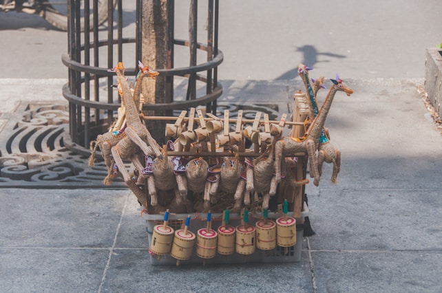 Handcrafted toys made of natural materials such as wood and straw are arranged on a street. The toys include colorful giraffe figures and cylindrical percussion instruments, displayed on a stone sidewalk near a tree.