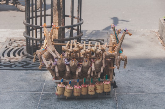 Handcrafted toys made of natural materials such as wood and straw are arranged on a street. The toys include colorful giraffe figures and cylindrical percussion instruments, displayed on a stone sidewalk near a tree.