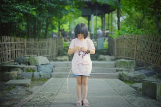 A young child stands on a stone pathway bordered by bamboo fencing, surrounded by lush greenery. The child is focused on an object in their hands and is wearing a light pink shirt and sandals.