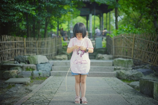 A young child stands on a stone pathway bordered by bamboo fencing, surrounded by lush greenery. The child is focused on an object in their hands and is wearing a light pink shirt and sandals.