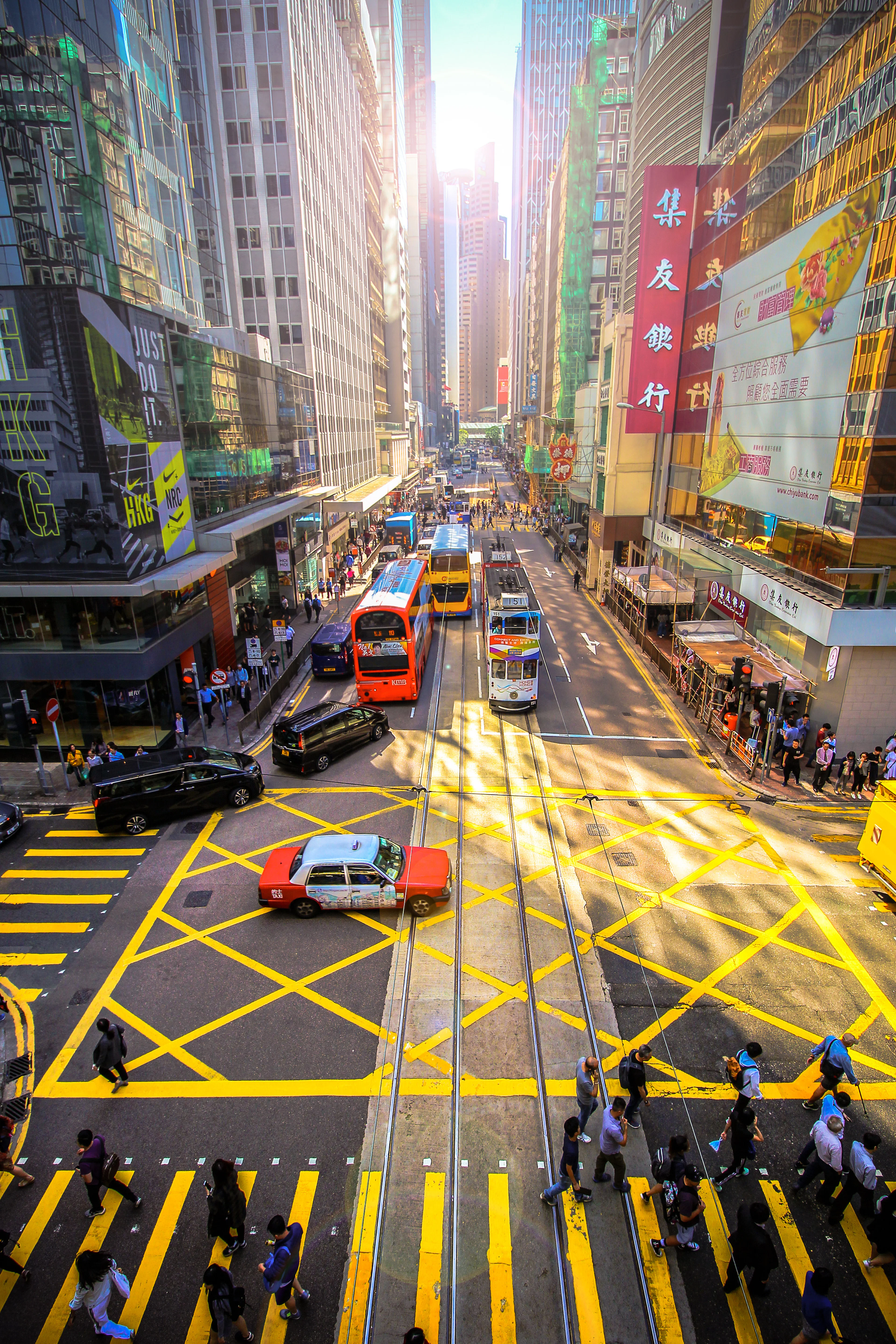A busy Hong Kong street crossing in low sun, with tram tracks and yellow box markings on the road, a red taxi and minibus in traffic, and Chinese shop signs on the towers above.