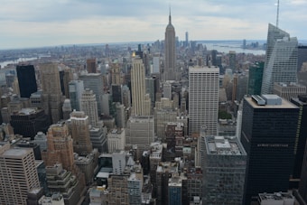 An extensive cityscape featuring numerous tall buildings and skyscrapers with the Empire State Building prominently visible in the center. The horizon shows more city buildings extending towards a body of water.