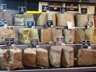 Various blocks of halva are displayed on shelves, each labeled with price tags. The halva appears in different flavors, indicated by tags with both Hebrew and English writing. A bright yellow knife is resting on the middle shelf.