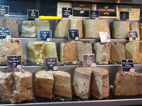 Various blocks of halva are displayed on shelves, each labeled with price tags. The halva appears in different flavors, indicated by tags with both Hebrew and English writing. A bright yellow knife is resting on the middle shelf.