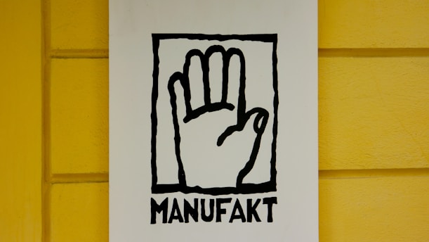 A stylized black outline of a hand inside a rectangular frame is centered on a white background. Below the hand, the word 'MANUFAKT' is written in bold, black letters. The background wall is painted in a bright yellow color with a subtle texture.
