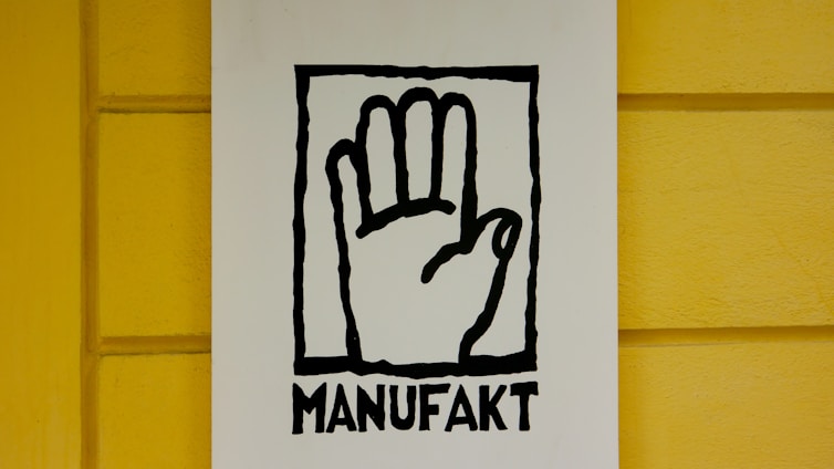 A stylized black outline of a hand inside a rectangular frame is centered on a white background. Below the hand, the word 'MANUFAKT' is written in bold, black letters. The background wall is painted in a bright yellow color with a subtle texture.