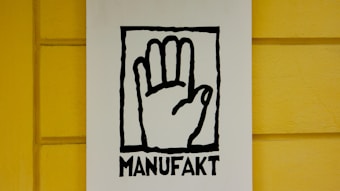 A stylized black outline of a hand inside a rectangular frame is centered on a white background. Below the hand, the word 'MANUFAKT' is written in bold, black letters. The background wall is painted in a bright yellow color with a subtle texture.
