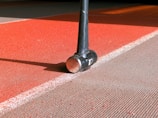 A close-up view of a black pole or handle placed on a textured athletic track with red and gray sections. The surface appears rough and is marked with a white line, casting a shadow from the pole.