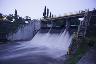 A concrete dam with a strong flow of water cascading down into a reservoir. The dam is bordered by stairways with blue and yellow railings on either side. Trees and greenery are visible in the background, and the sky is overcast, suggesting an evening or early morning setting.