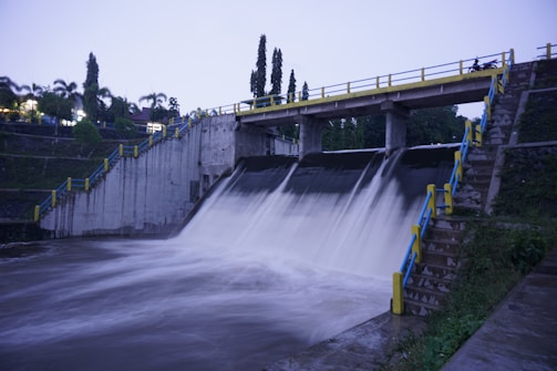 A concrete dam with a strong flow of water cascading down into a reservoir. The dam is bordered by stairways with blue and yellow railings on either side. Trees and greenery are visible in the background, and the sky is overcast, suggesting an evening or early morning setting.