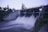 A concrete dam with a strong flow of water cascading down into a reservoir. The dam is bordered by stairways with blue and yellow railings on either side. Trees and greenery are visible in the background, and the sky is overcast, suggesting an evening or early morning setting.